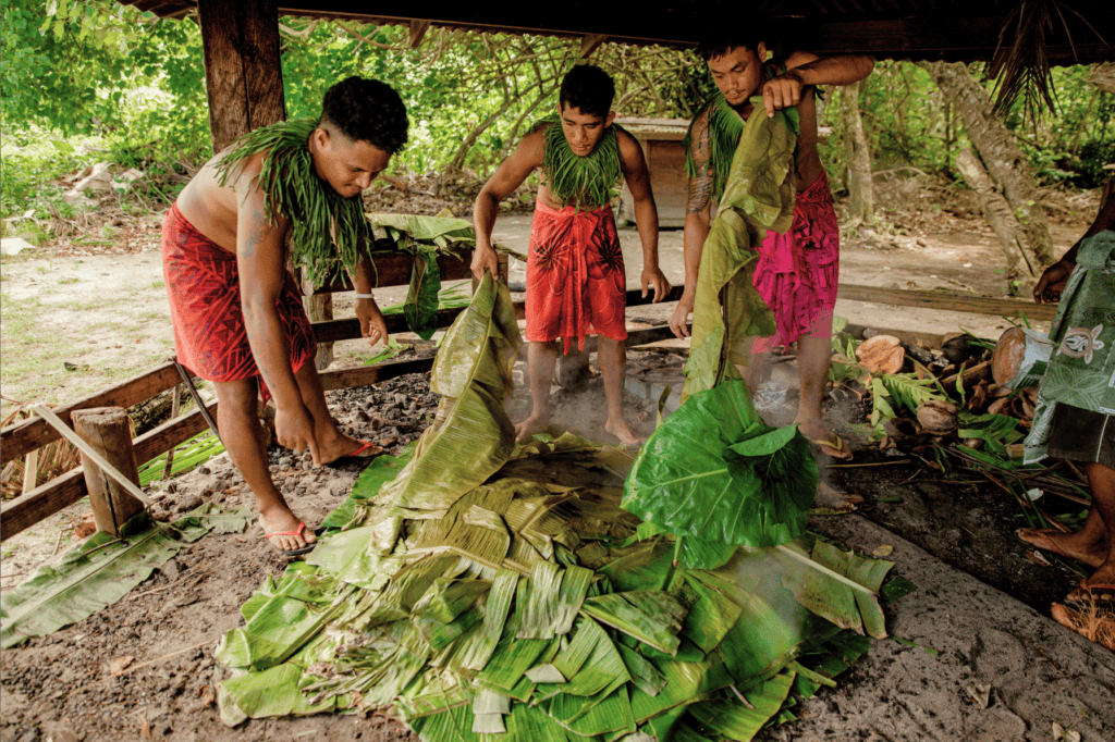 Samoan men take leaves off food cooking in umu.